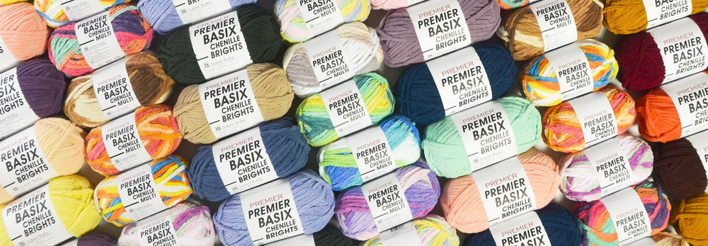 A colorful assortment of Premier Basix Chenille yarn skeins arranged in a diagonal pattern. The skeins come in a variety of solid and multicolored shades, including bright, pastel, and neutral tones. Each skein is wrapped with a white label - Premier Basix Chenille