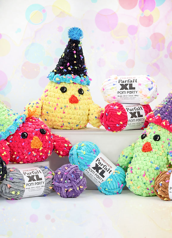 A colorful display of plush, crocheted bird toys made from Premier Parfait XL Pom Party yarn. The birds are crafted in bright hues like red, yellow, green, and purple, each wearing a playful, cone-shaped party hat with pom-pom tips. Surrounding the birds are several skeins of the plush, multicolored yarn in shades of black, gray, yellow, purple, blue, and brown. The background features a festive pastel pattern with bubbles and marbled textures.